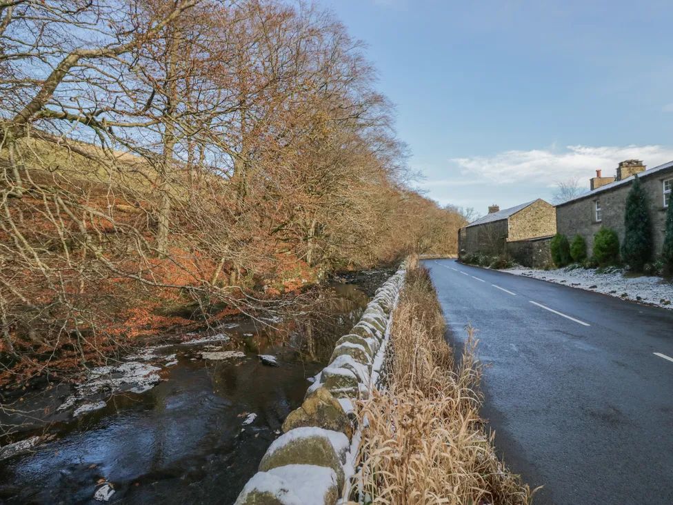 A narrow road alongside a stone wall with a stream and leafless trees on the left and stone buildings on the right at The Old Cart House in Garsdale near Sedbergh