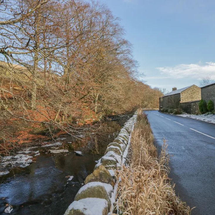A snowy winter lane beside a stone wall and beck, with stone cottages near The Old Cart House