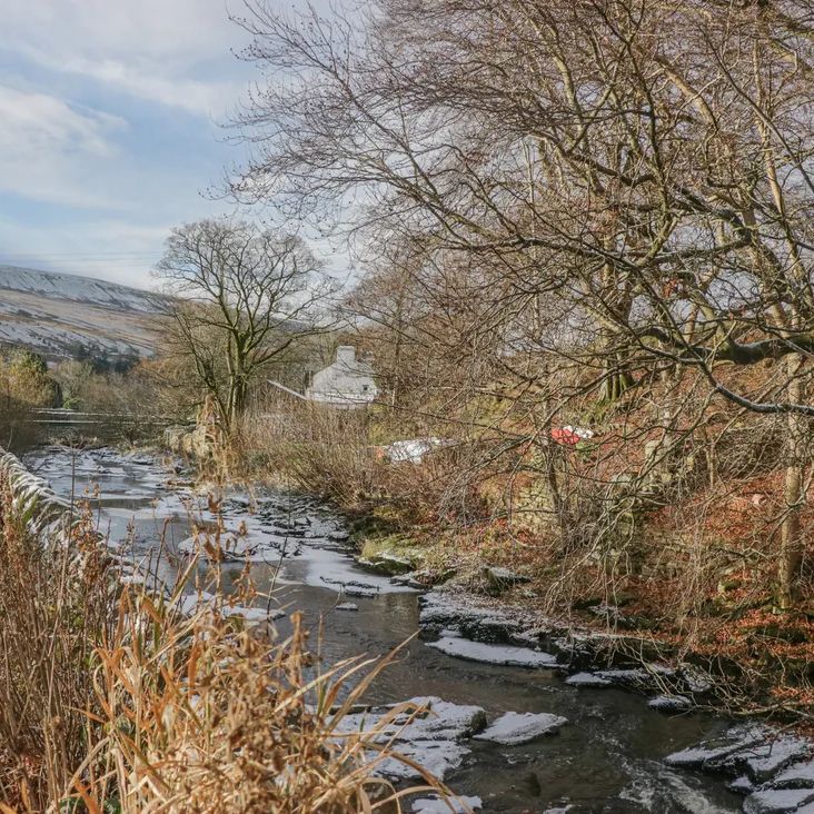 A winter beck flowing past a road and bare trees, with a stone cottage near The Old Cart House