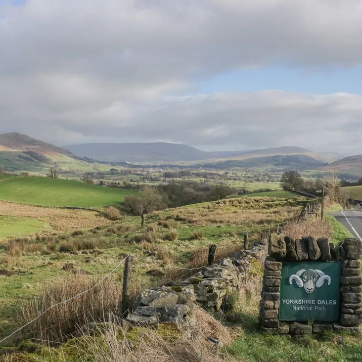 A Yorkshire Dales National Park sign set in a stone wall beside a road, with fields and fells beyond