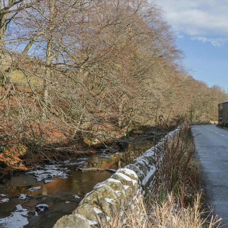 A sunny winter lane beside a snow-topped dry-stone wall and beck, with a stone building near The Old Cart House