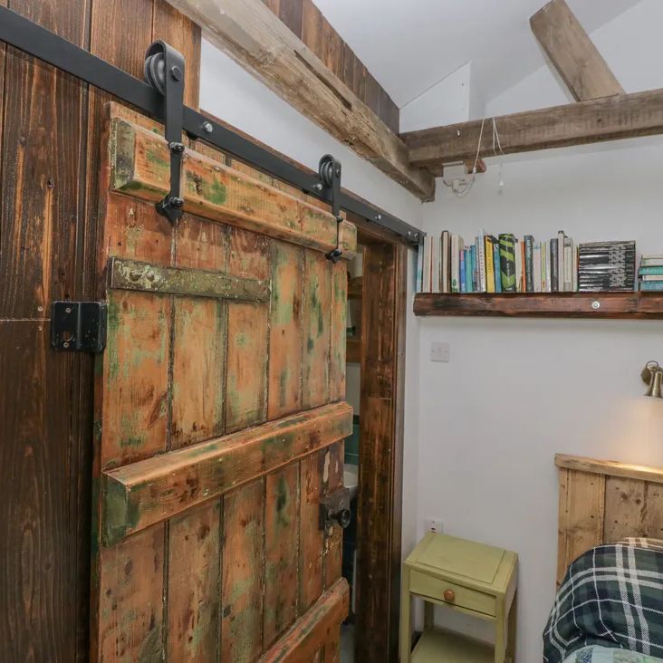 A rustic sliding barn door beside a bed and book shelf in a beamed bedroom at The Old Cart House
