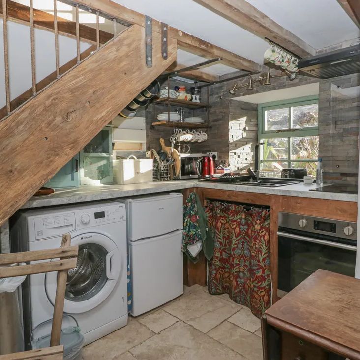 The compact kitchen at The Old Cart House with a washing machine under the stairs, open shelving and a green-framed window