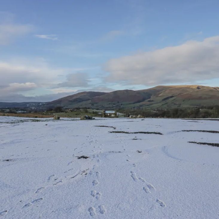 A snowy field with footprints leading to a gate, with Dales hills beyond, near The Old Cart House