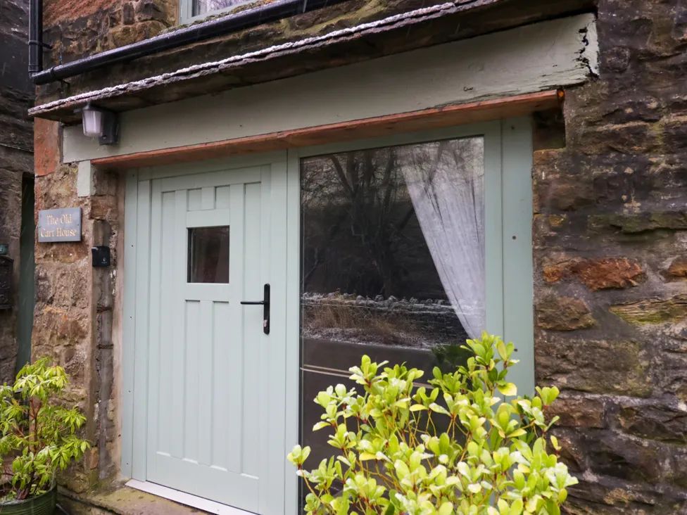 A stone building entrance with a light green door and window with curtains and plants near the door at The Old Cart House in Garsdale near Sedbergh