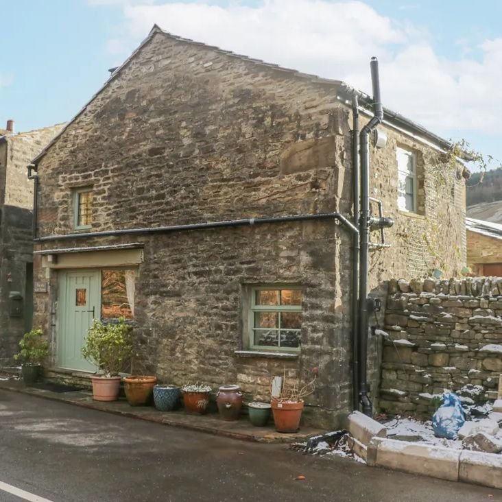 The stone exterior of The Old Cart House with sage-green woodwork and potted plants, beside a snowy lane