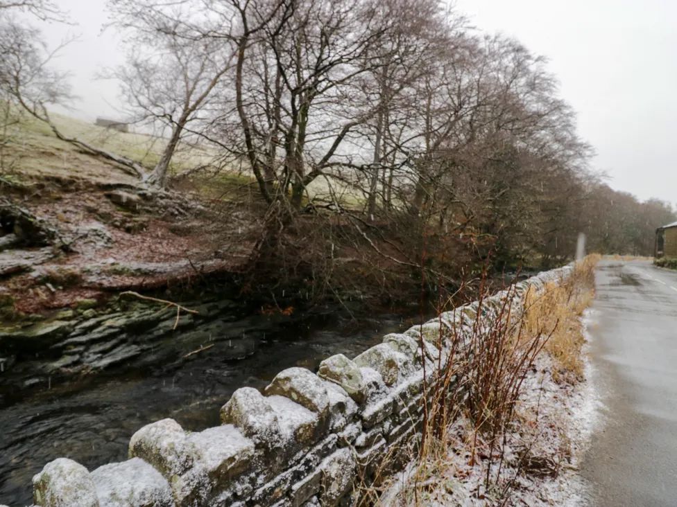 A stone wall alongside a narrow road next to a river with leafless trees and light snow at The Old Cart House in Garsdale near Sedbergh