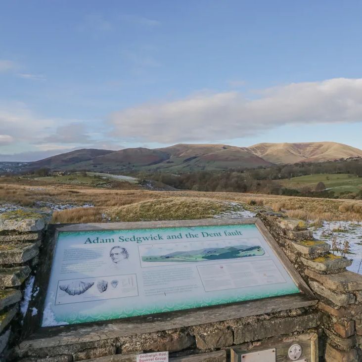 An interpretation board about Adam Sedgwick and the Dent Fault set in a dry-stone wall, with snowy Dales hills beyond