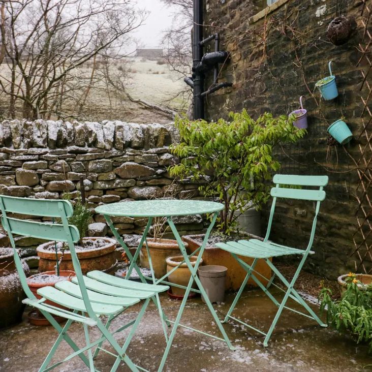 A snow-dusted patio at The Old Cart House with a mint-green bistro set and potted plants by a stone wall