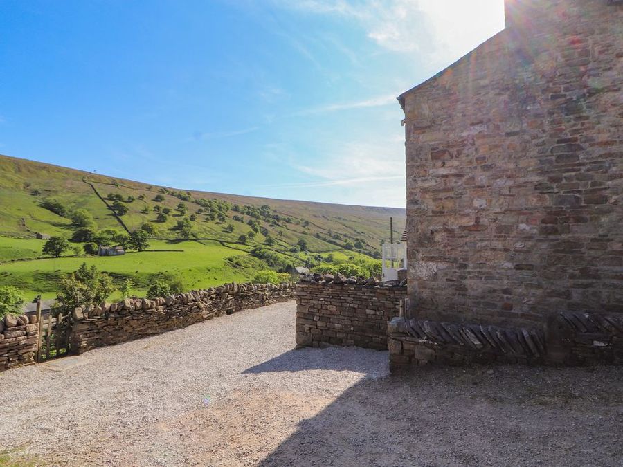 Stone wall and gravel path beside a stone building with green hills and trees in the background at Roger Pot in Garsdale near Sedbergh